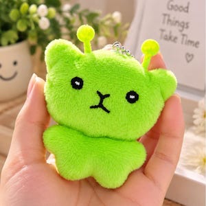 Peut inclure: Un porte-clés en peluche vert citron vif en forme de créature stylisée avec deux antennes, des yeux noirs ronds et une bouche noire brodée. Ce personnage doux et duveteux possède une petite chaînette à billes en métal fixée sur le dessus.