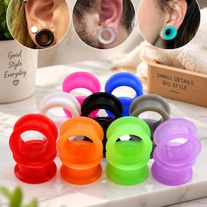Peut inclure: Une collection de tunnels d'oreille en silicone de différentes couleurs unies, notamment rouge, orange, vert néon, violet, blanc, noir, bleu et gris. Les tunnels sont circulaires avec un bord évasé, disposés en groupe sur une surface en marbre blanc.