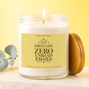 May include: A white soy wax candle in a clear glass jar with a gold lid. The yellow label features the text DUSKANDFLAME, an envelope icon, and the words SMELLS LIKE ZERO UNREAD EMAILS, with %100 SOY WAX CANDLE | ECO-FRIENDLY | HANDPOURED below.