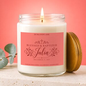 May include: A clear glass jar candle with a lit wick and a coral-colored label. The label features the text DUSKANDFLAME, BLESSED & BAPTIZED, Julia, and DECEMBER 17, 2027, with a cross and floral graphics. A gold-colored metal lid sits beside the jar.
