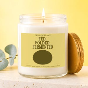 May include: A white soy wax candle in a clear glass jar with a gold lid. The yellow label features the text DUSKANDFLAME, FED, FOLDED, FERMENTED, and %100 SOY WAX CANDLE | ECO-FRIENDLY | HANDPOURED, along with a dark olive green oval graphic.