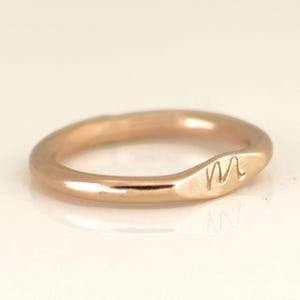 May include: A rose gold ring with a smooth, rounded band. The ring features a slightly flattened oval design with the letter "M" engraved on the surface. The ring is displayed on a white surface.