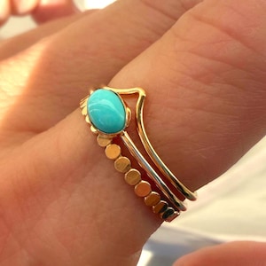 May include: A set of three gold rings. One ring features a turquoise oval stone, another has a curved design, and the third has a series of small gold circles. The rings are stacked together.