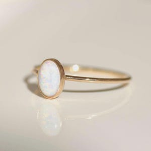 May include: A gold ring with an oval opal gemstone. The ring has a slender band and a bezel setting. The opal displays a range of colours. The ring is shown on a reflective surface. The design is elegant and simple.