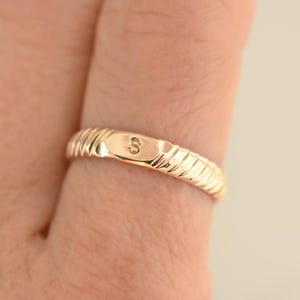 May include: A gold-colored ring with a ribbed band design. The ring features a small, rectangular plate with the letter "S" engraved on it. The ring is shown on a finger.
