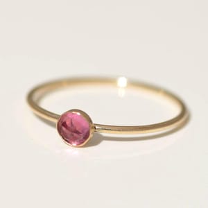 Puede incluir: Un delicado anillo de oro con una gema rosa vibrante y redonda. La banda del anillo es fina y pulida, creando un diseño minimalista y elegante. La gema es el punto focal, engastada en un bisel simple.