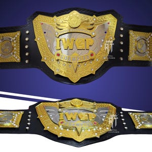 Könnte beinhalten: Ein schwarzer Leder-Meisterschaftsgürtel mit einer großen, verzierten Gold- und Silber-Mittelplatte mit der Aufschrift IWGP WORLD HEAVYWEIGHT CHAMPION. Die Platte ist mit kleinen roten und blauen Edelsteinen und Seitenplatten mit Weltkarten verziert.