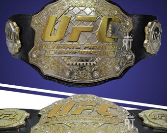 May include: A black leather championship belt featuring a large central metal plate with gold UFC lettering and the text Ultimate Fighting Championship. The plate has a silver filigree border with embedded clear stones and two smaller side plates.