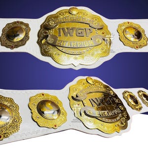 May include: A white leather championship belt featuring a large, ornate gold-plated center plate with the text INTERCONTINENTAL IWGP CHAMPION. The belt includes smaller gold side plates with red accents and intricate engraved detailing throughout.