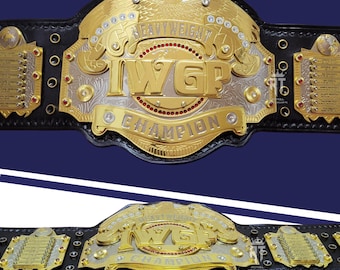 Pode incluir: Um cinturão de campeonato em couro preto com uma grande placa central dourada apresentando um emblema de águia, o texto IWGP HEAVYWEIGHT CHAMPION e pedras vermelhas e transparentes. Duas placas laterais exibem nomes gravados.