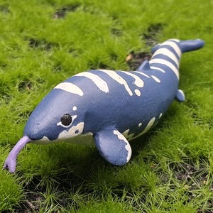 May include: A blue and white toy narwhal figurine with a purple tongue, lying on a bed of green artificial grass. The narwhal has white markings and a black eye. The figurine is a whimsical representation of a marine mammal.