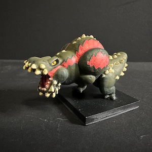May include: A detailed figurine of a cartoonish monster, primarily in shades of green and red, with yellow accents. The creature has an open mouth revealing teeth, and is mounted on a black base.