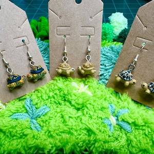 May include: Three pairs of dangle earrings featuring miniature turtle charms. The turtles are painted in shades of brown, black, and yellow. The earrings are displayed on brown card stock.