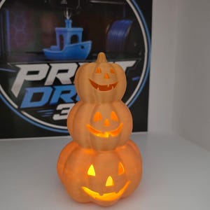 May include: A stack of three orange jack-o'-lantern pumpkins with carved triangular eyes and smiling mouths. The pumpkins are illuminated from within, casting a warm glow through the cutouts. The structure is vertically oriented with a stem on top.