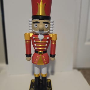 Può includere: Una statuetta schiaccianoci che indossa un'uniforme rossa con dettagli dorati, un alto cappello rosso e capelli bianchi. La figura poggia su una base rettangolare nera con disegni di fiocchi di neve dorati e la scritta Merry Xmas in oro.