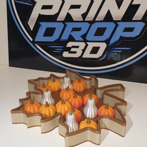May include: A maple leaf-shaped tray with tan walls and a brown rim, containing a collection of small, 3D-printed orange pumpkins and silver-colored gourds. The background features a sign with the text PRINT DROP 3D.