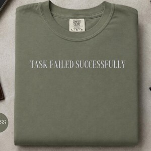 Puede incluir: Una camiseta de cuello redondo color verde musgo con el texto blanco "TASK FAILED SUCCESSFULLY" impreso en el pecho. La prenda tiene una etiqueta de la marca Comfort Colors en el cuello interior con la talla L. La tela tiene un acabado mate.