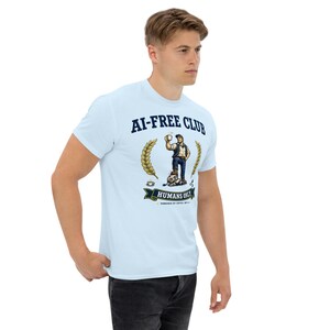 May include: A light blue t-shirt featuring a graphic with the text AI-FREE CLUB and HUMANS ONLY. The design shows a person holding a coffee cup, standing over a robot, framed by wheat stalks. The text also includes POWERED BY COFFEE, NOT AI.