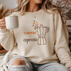 May include: A cream-colored crewneck sweatshirt featuring a brown line-art illustration of a moka pot with steam, coffee beans, and orange pumpkin icons. The text reads PUMPKINS & espresso in a mix of orange and brown fonts on the front chest.