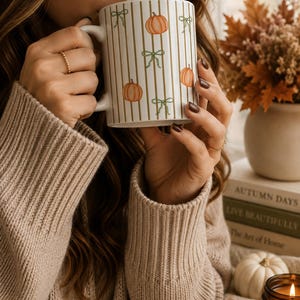 Peut inclure: Un mug en céramique blanche orné d'un motif de rayures verticales dorées, de citrouilles orange et de nœuds vert clair. Le mug présente un corps cylindrique lisse et une anse incurvée, affichant un design saisonnier aux illustrations nettes.