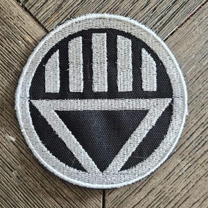 May include: A circular embroidered patch featuring a black background with a white border. The design consists of a white stylized hand with four vertical fingers above a large white downward-pointing triangle centered within the circle.