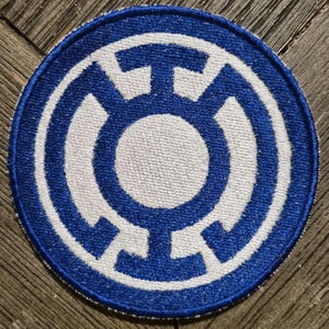 May include: A circular embroidered patch featuring a blue and white design. The emblem consists of a central white circle surrounded by a blue ring, with four symmetrical blue extensions reaching outward toward the edge of the circular border.