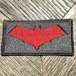 May include: A rectangular embroidered patch featuring a black background with a centered, vibrant red bat-shaped emblem. The patch has a thick black stitched border, and the entire surface displays a consistent, dense thread texture throughout the design.