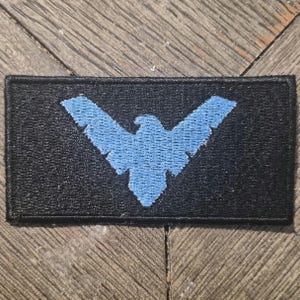 May include: A rectangular embroidered patch featuring a black background with a light blue bird emblem centered on the fabric. The bird design has outstretched wings and a detailed silhouette, with a stitched border surrounding the entire patch.