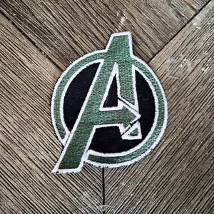 Peut inclure: Un écusson brodé circulaire représentant la lettre A dans une police stylisée. La lettre est vert foncé avec une bordure blanche, sur un fond circulaire noir uni. L'ensemble du motif est souligné par une couture blanche sur une base en tissu.