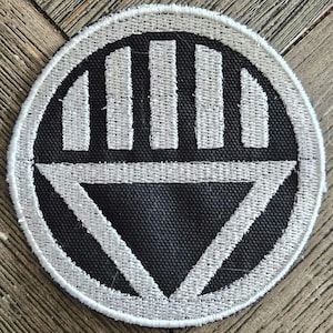 May include: A circular embroidered patch featuring a black background with a white border and a central white graphic design. The design consists of a horizontal bar above a downward-pointing triangle, with four vertical bars positioned above the horizontal line.