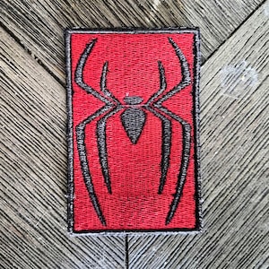 May include: A rectangular embroidered patch featuring a vibrant red background with a black stitched spider emblem centred on the surface. The patch has a thick black border outlining the rectangular shape and the spider design is detailed with thin lines.