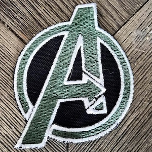 May include: A circular embroidered patch featuring the Avengers logo. The design consists of a large letter A in a metallic olive green thread, outlined in white, set against a solid black circular background with a white stitched border.