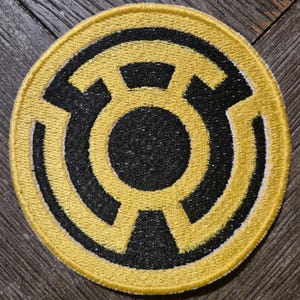 May include: A circular embroidered patch featuring a yellow and black design. The emblem consists of a central black circle surrounded by a yellow ring, with four yellow segments extending outward to form a stylized lantern-like symbol on a black background.