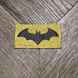 May include: A rectangular embroidered patch featuring a black bat silhouette centred on a yellow background. The patch has a stitched border and a textured fabric surface, displaying a classic winged emblem design in a high-contrast colour scheme.