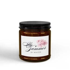 Peut inclure: Un pot à bougie cylindrique en verre ambré avec un couvercle noir à vis. L'étiquette rectangulaire blanche présente un bouquet de fleurs d'hortensia aquarelle rose clair et le texte noir "Ily Summer BY WINTER" en polices script et sans-serif.