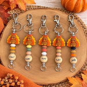 May include: Five turkey-themed keychains featuring silicone turkey charms with orange and yellow feathers, silver lobster clasps, and wooden beads. Each keychain includes a bead printed with the words blessed, grateful, harvest, thankful, or gather.