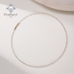 May include: A delicate necklace featuring a continuous strand of small, round white pearls. The necklace includes a single, smooth, cylindrical gold-toned metal clasp component positioned on the left side of the circular arrangement.