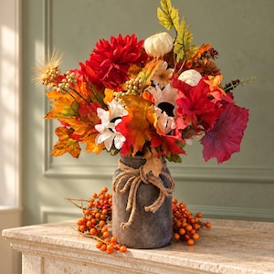 May include: A floral arrangement featuring red dahlias, white sunflowers, small white pumpkins, and autumn-colored leaves in a dark gray cylindrical vase wrapped with a thick jute rope bow, accented by orange berry sprigs at the base.