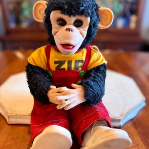 May include: A plush monkey doll with black fur, a tan face, and large dark eyes. It wears a yellow t-shirt with the word ZIP printed in black, red corduroy overalls with green buttons, and white shoes. The doll is seated with hands clasped together.