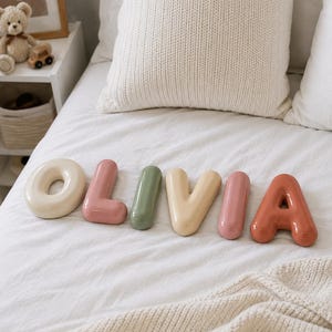 May include: A set of six individual, smooth, rounded, three-dimensional letters spelling the name OLIVIA. The letters are finished in a glossy, multi-colored palette including cream, dusty pink, sage green, light yellow, and terracotta orange.