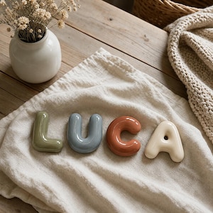 May include: Four individual ceramic letters spelling the name LUCA. The letter L is sage green, the letter U is slate blue, the letter C is terracotta orange, and the letter A is cream. Each letter has a smooth, rounded, and glossy finish.