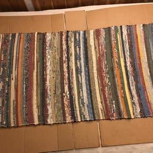 May include: A rectangular rag rug featuring a multi-colored striped pattern with various woven fabric strips in shades of green, blue, red, yellow, and white. The rug has a textured, woven construction and includes white fringe along both short ends.