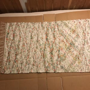 May include: A rectangular woven rag rug featuring a predominantly white base with interspersed flecks of green, orange, and yellow fabric strips. The rug has a textured, multi-colored appearance and includes decorative fringe along both short ends.