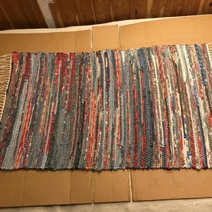 May include: A rectangular rag rug featuring a woven pattern of multicolored fabric strips in shades of blue, red, grey, and white. The rug has a textured, multi-tonal appearance with visible frayed edges and off-white fringe tassels on both ends.