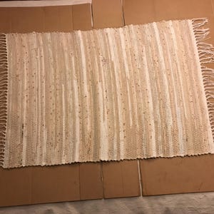 May include: A rectangular woven rag rug featuring a striped pattern in shades of cream, beige, and light brown. The rug is constructed with tightly woven fabric strips and finished with long, knotted fringe detailing along both shorter ends.