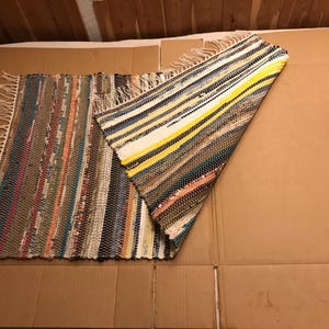 May include: A rectangular woven rag rug featuring a multicolored striped pattern with hues of yellow, blue, green, and brown. The rug has a folded corner revealing the underside and is finished with off-white fringe along the short edges.