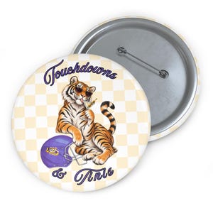 May include: A round pin-back button featuring a cream and white checkered background. A tiger wearing sunglasses holds a martini glass, resting next to a purple football helmet. The text reads Touchdowns & Tinis in purple script lettering.