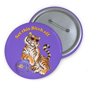 May include: A round purple pin-back button featuring a tiger wearing sunglasses, holding a martini glass with olives, and resting its paw on a purple football helmet. The text "Set this Bitch off" is printed in yellow letters above the tiger.