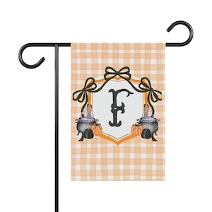 Peut inclure: Un drapeau de jardin rectangulaire avec un motif vichy orange clair et blanc. Le centre présente un écusson blanc avec une lettre F noire, encadré par des nœuds noirs, deux chaudrons noirs avec des jambes rayées, et des citrouilles orange et noires.
