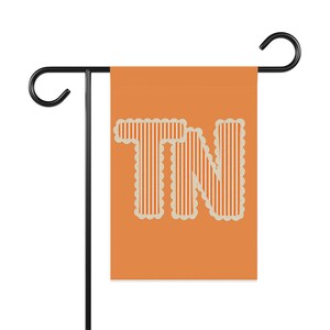 May include: A rectangular garden flag in a solid orange color featuring the letters TN in a white and orange vertical striped pattern. The letters are outlined with a white scalloped border and the flag hangs from a black metal stand.